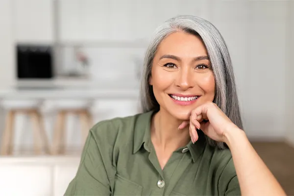 Menopause-Clinic An asian woman with long gray hair and a green shirt sitting and smiling on her couch, happy with her menopause care from [NAME} in Northern New Jersey.