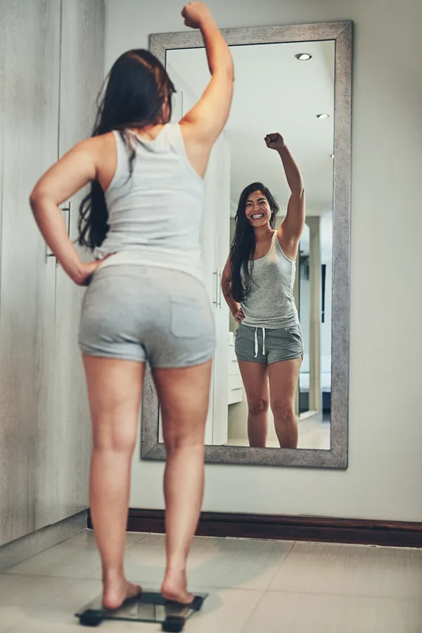 Tirzepatide-Treatment A woman in workout clothes stands on a scale looking in the mirror and pumping her hands in celebration of her successful Tirzepatide treatment from Healthy Aging Medical Centers of New Jersey in Northern New Jersey.